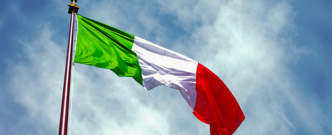 Italian flag waving on a tall flagpole against a partly cloudy blue sky.