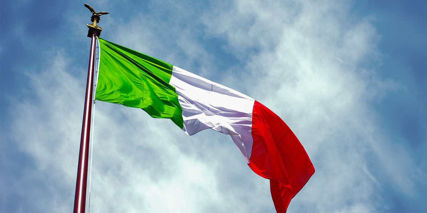 Italian flag waving on a tall flagpole against a partly cloudy blue sky.