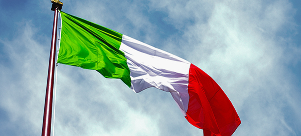 Italian flag waving on a tall flagpole against a partly cloudy blue sky.