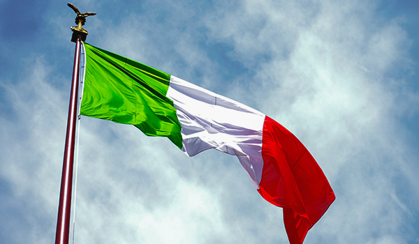 Italian flag waving on a tall flagpole against a partly cloudy blue sky.